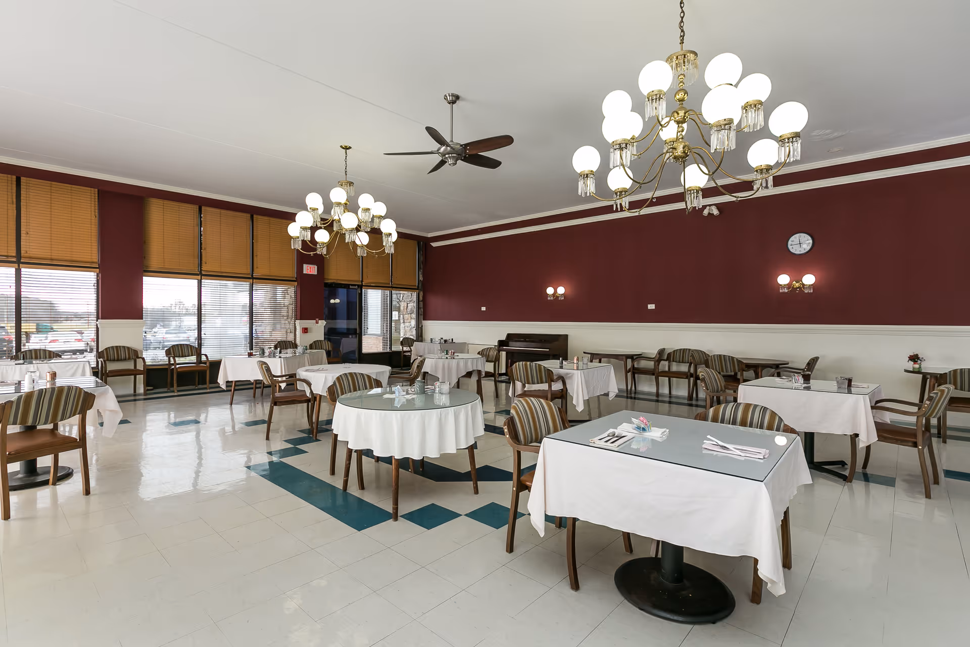 A spacious dining room with multiple tables covered in white tablecloths, each set with utensils and napkins. The room features striped cushioned chairs, large windows with blinds, maroon and white walls, two chandeliers, ceiling fans, and a clock on the wall.