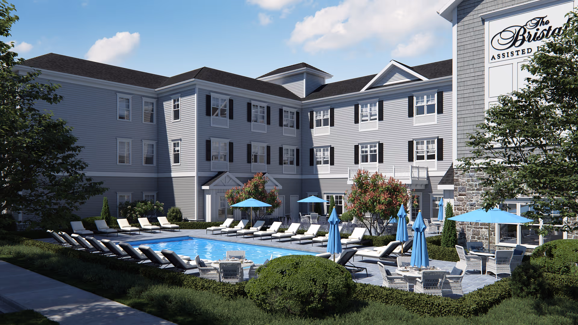 Outdoor view of The Bristal Assisted Living at Somerset featuring a swimming pool surrounded by lounge chairs and tables with blue umbrellas, set against a three-story building with gray siding and white trim under a blue sky with some clouds.
