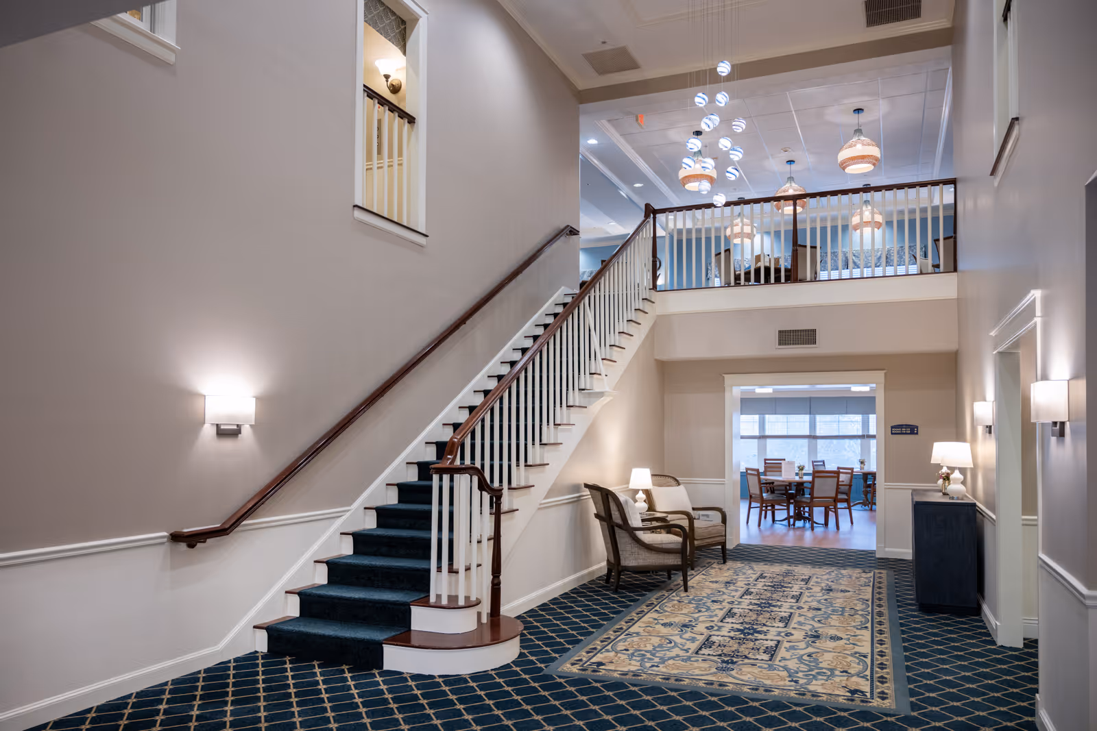 Interior view of Stafford Hill Assisted Living showing a carpeted staircase with wooden handrails leading to an upper level. The hallway features patterned carpeting, two cushioned chairs with a small table and lamp between them, wall sconces providing soft lighting, and a glimpse into a dining area with tables and chairs in the background. Modern pendant lights hang from the ceiling above the upper level.