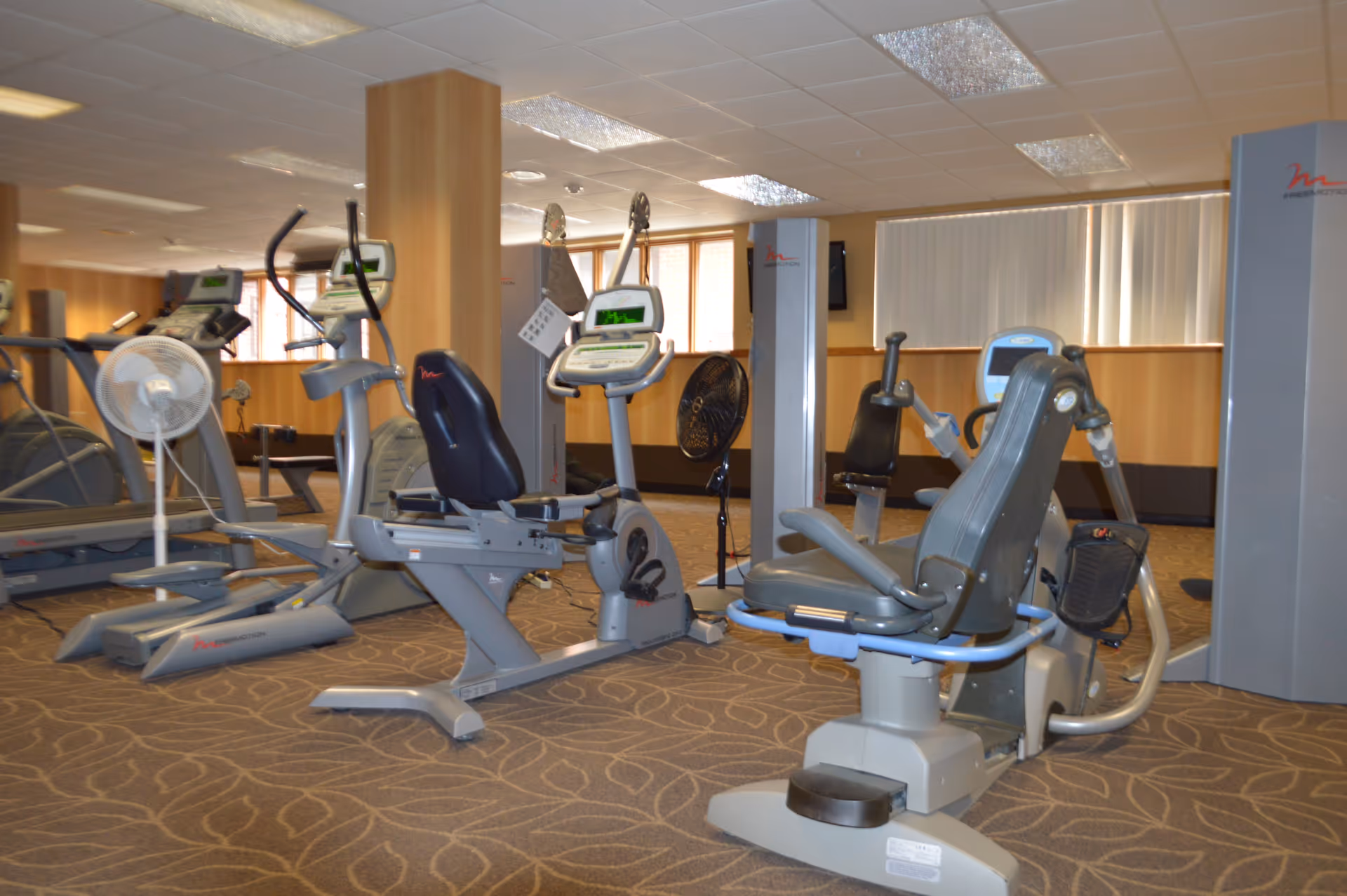 Interior view of a fitness room in a senior living facility with various exercise machines including stationary bikes, elliptical trainers, and weight machines. The room has carpeted flooring with a leaf pattern, wood-paneled walls, and large windows with vertical blinds.