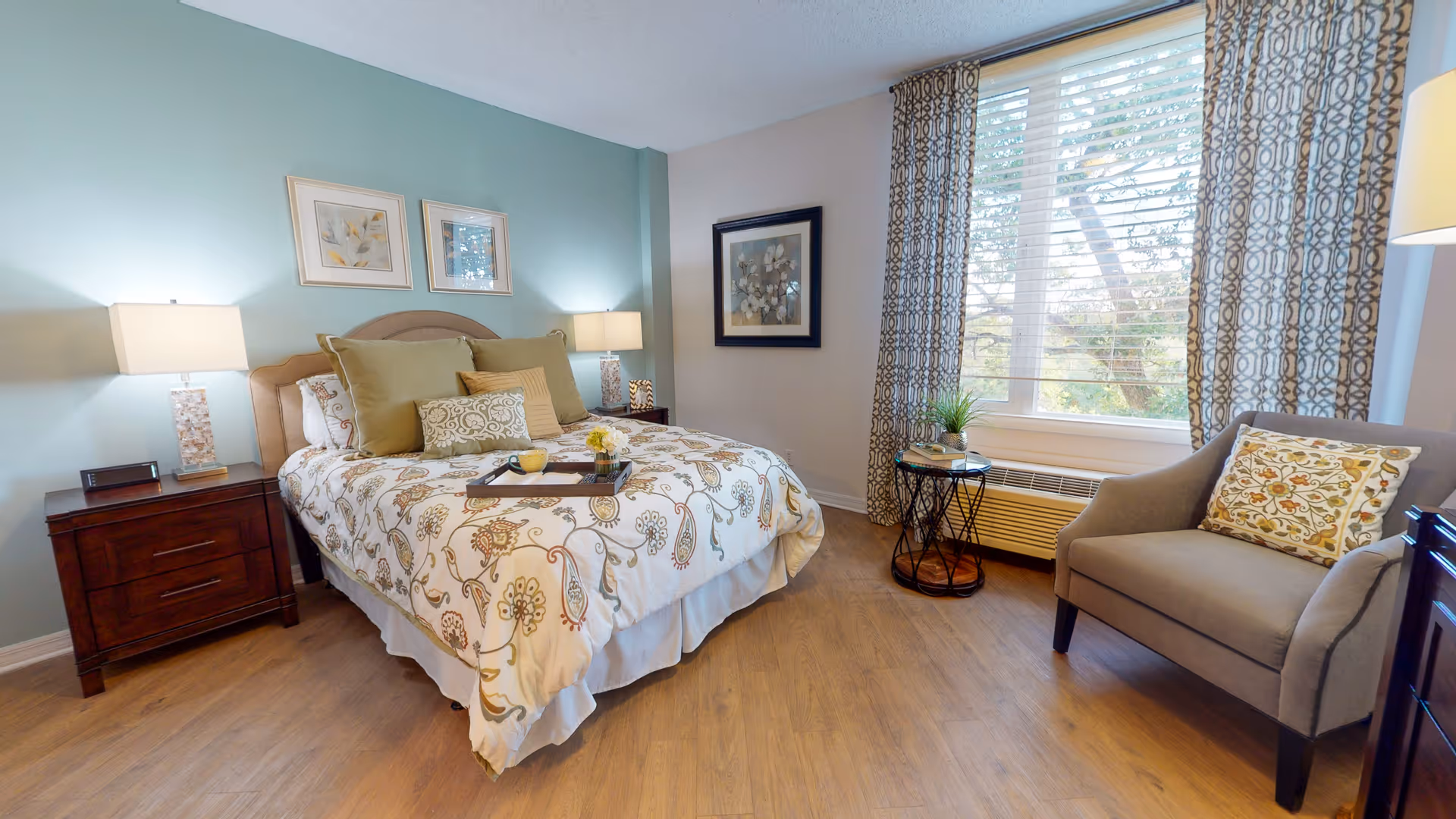 A cozy bedroom with a bed featuring floral patterned bedding and multiple pillows. There are two bedside tables with lamps on either side of the bed. A framed picture hangs on the wall above the bed, and another framed artwork is on the adjacent wall. A large window with patterned curtains lets in natural light. A comfortable armchair with a decorative pillow and a small round side table with a plant and books are placed near the window. The room has wooden flooring and a calm, inviting atmosphere.