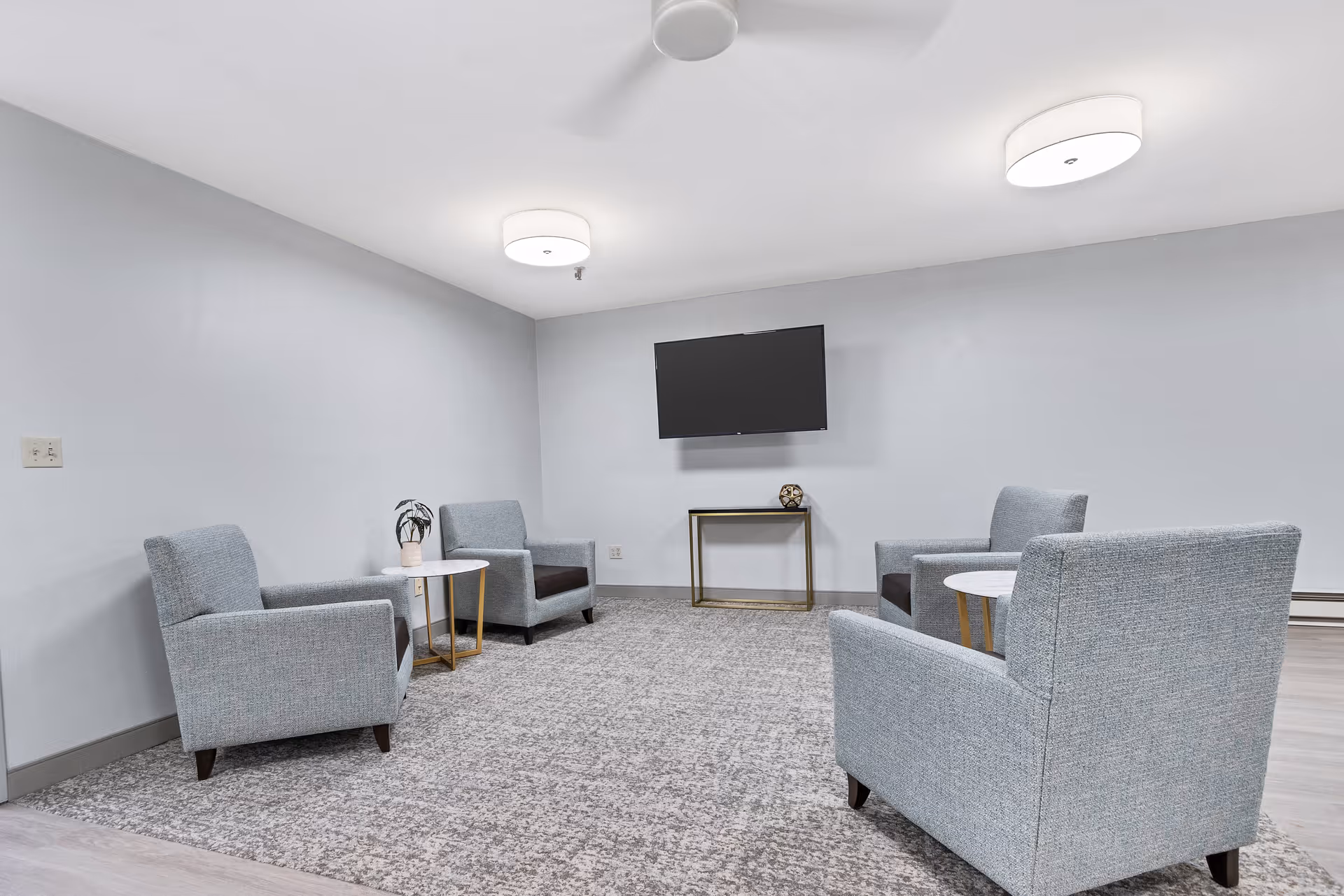 A small sitting lounge with four gray armchairs arranged around side tables facing a wall-mounted TV.