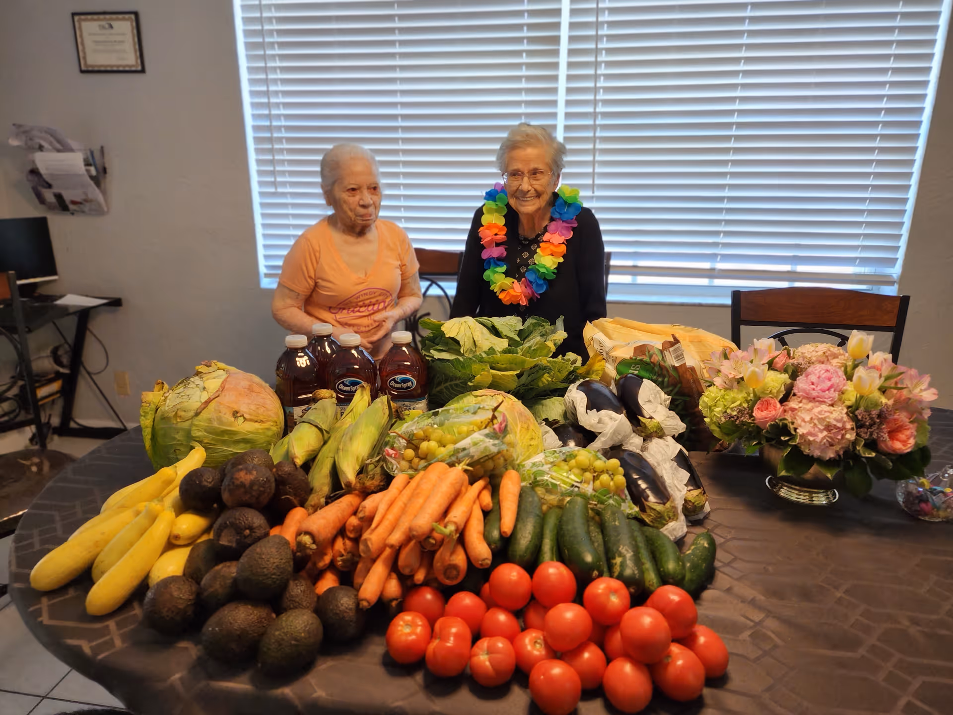 Two elderly women sitting at a table indoors with a large assortment of fresh vegetables and fruits including bananas, avocados, carrots, tomatoes, cucumbers, corn, cabbage, and grapes. One woman is wearing an orange shirt and the other is wearing a black top with a colorful flower lei. There is also a bouquet of flowers on the table and a window with blinds in the background.