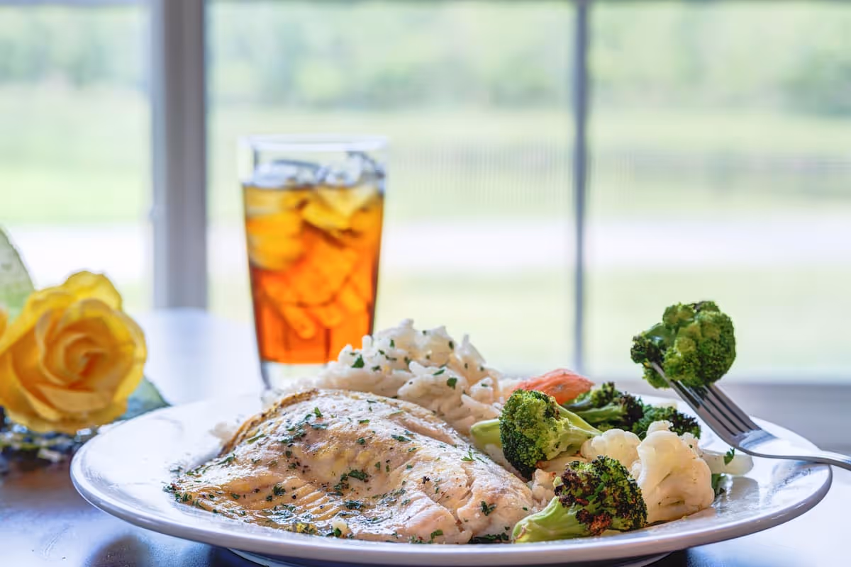 A plate of food with grilled fish, steamed broccoli and cauliflower, white rice, and a slice of tomato, accompanied by a glass of iced tea and a yellow rose on the side, set on a table near a window with a blurred outdoor background.