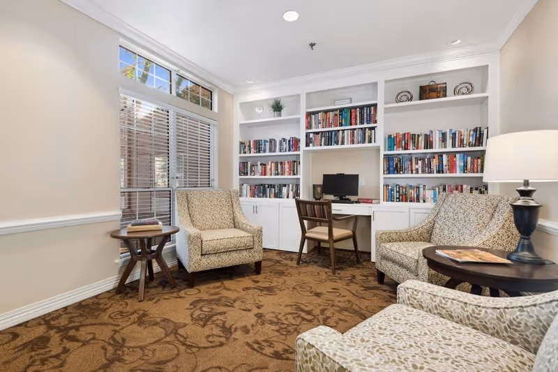 A cozy reading room with three patterned armchairs arranged around two small round wooden tables. One table has a book on it and the other has a lamp. A large built-in white bookshelf filled with books and decorative items is against the back wall, with a small desk and computer in the center. A window with blinds lets in natural light.