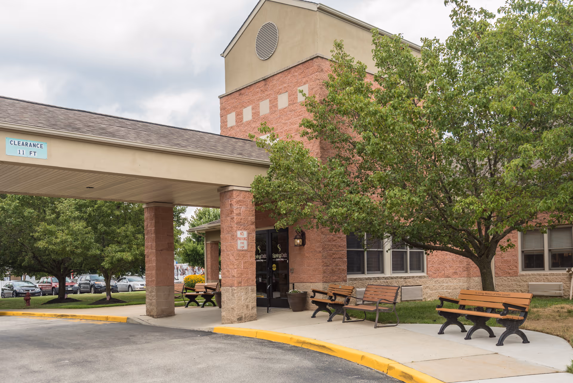 Entrance area of a senior living facility with a covered drop-off zone supported by brick columns. There are several wooden benches on the sidewalk near the entrance, and a large tree providing shade. The building has a brick and beige exterior with windows. A clearance sign indicating 11 feet is visible on the covered area.