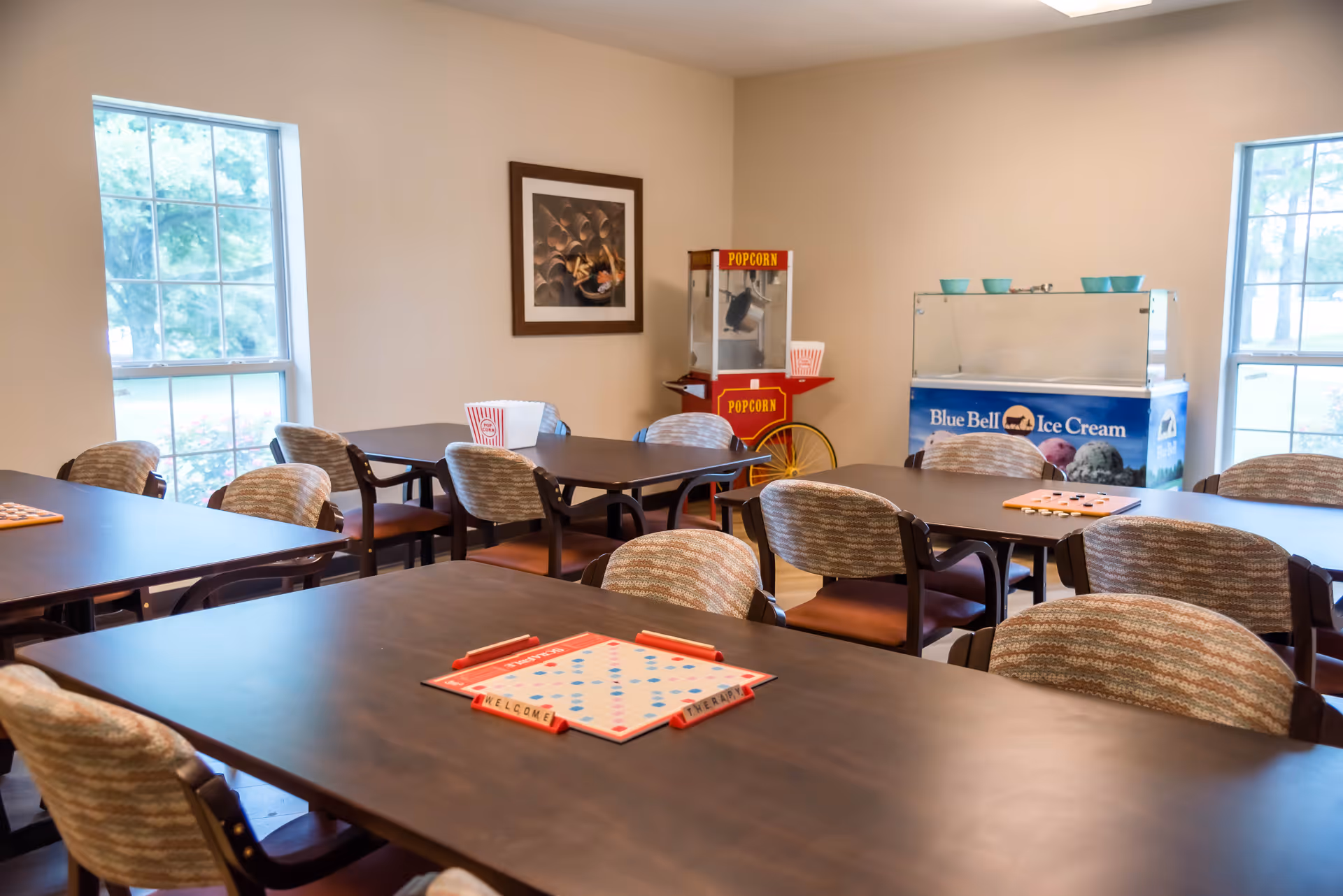 A bright communal dining/activity room with multiple tables and chairs, board games on the tables, a popcorn cart, and a Blue Bell ice cream freezer against the wall.