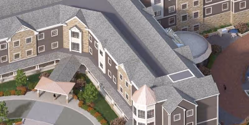 Aerial view of a senior living facility building with multiple floors, gray roofs, and brown and white exterior walls. The building surrounds a landscaped area with trees, shrubs, and a covered entrance driveway.