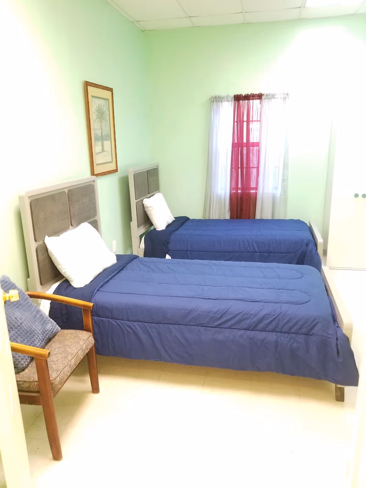 A small bedroom with two single beds side by side, each with a blue bedspread and a white pillow. There is a wooden chair with a cushion and a blue pillow on it near the foot of the beds. The walls are painted light green, and there is a framed picture of a palm tree on the wall. A window with white and red curtains is visible at the far end of the room.