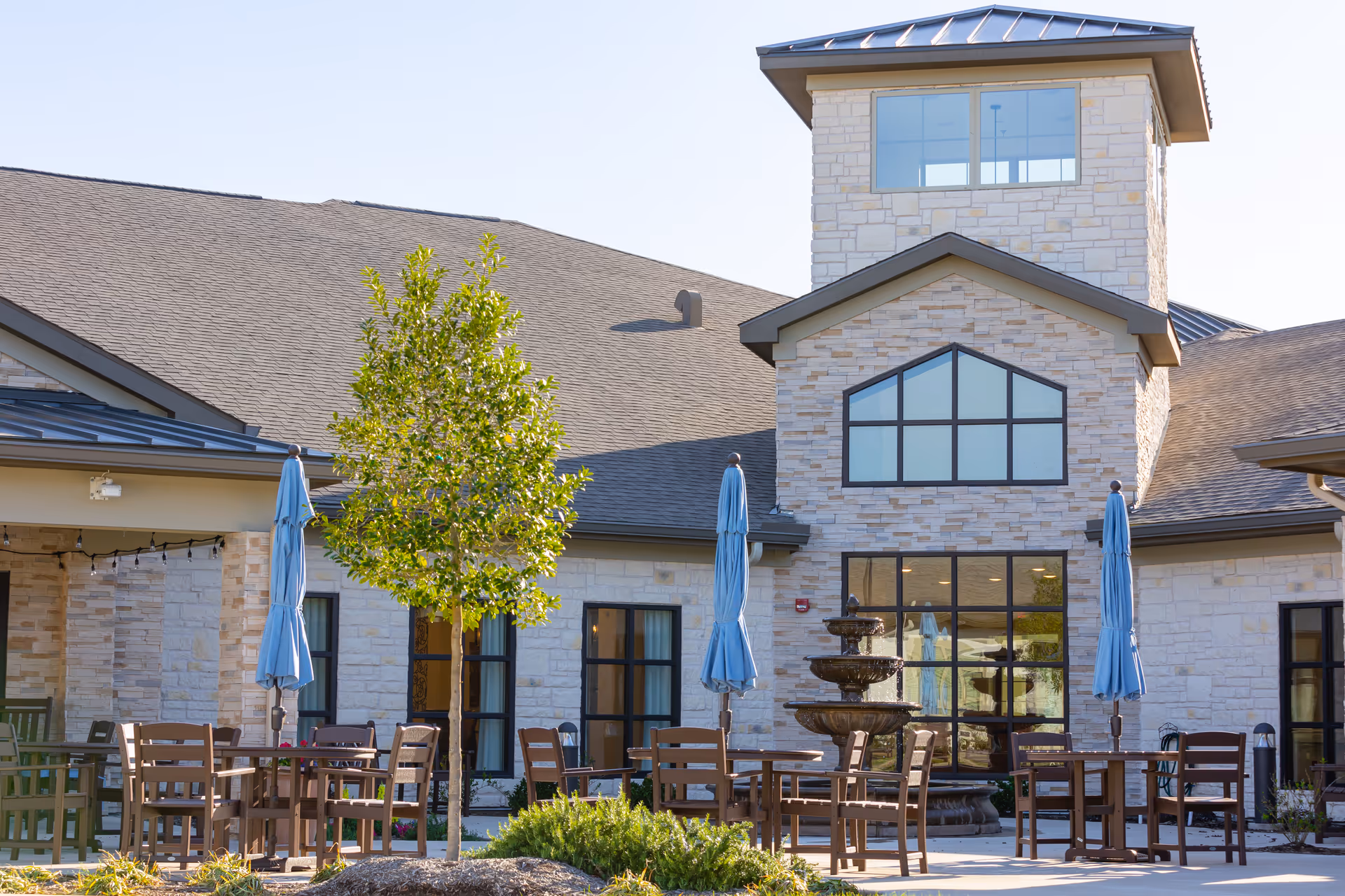 Outdoor patio area at The Village at Sugar Land featuring wooden tables and chairs with closed blue umbrellas, a small tree, and a multi-tiered water fountain in front of a stone building with large windows.