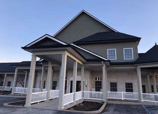 Exterior view of a senior living facility building with a covered entrance supported by columns, white railings, and multiple windows. The sky is clear and the building has a combination of brick and siding.