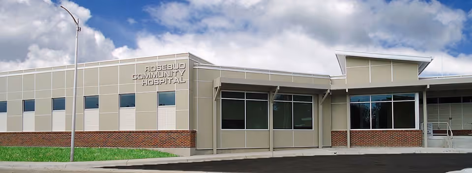 Exterior view of Rosebud Community Hospital building under a partly cloudy sky, featuring a modern design with large windows and a combination of beige panels and red brick accents.