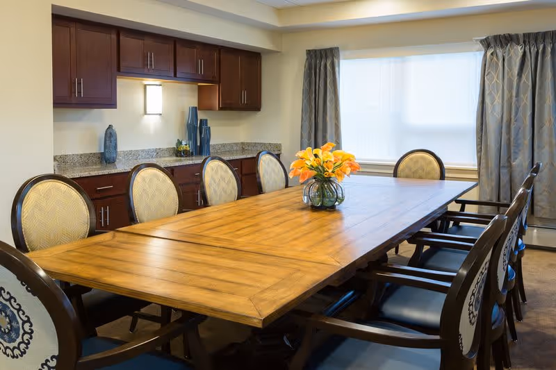 A well-lit dining room with a long wooden table surrounded by eight cushioned chairs. On the table is a glass vase with orange flowers. The room has a granite countertop with dark wooden cabinets above and below it. There is a window with patterned curtains allowing natural light into the room.