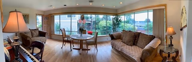 A bright living room with large windows overlooking a garden. The room features a brown sofa with cushions, a round table with two chairs, a piano with a lamp on it, and two side tables with lamps. The floor is wooden, and the walls are light-colored, creating a warm and inviting atmosphere.