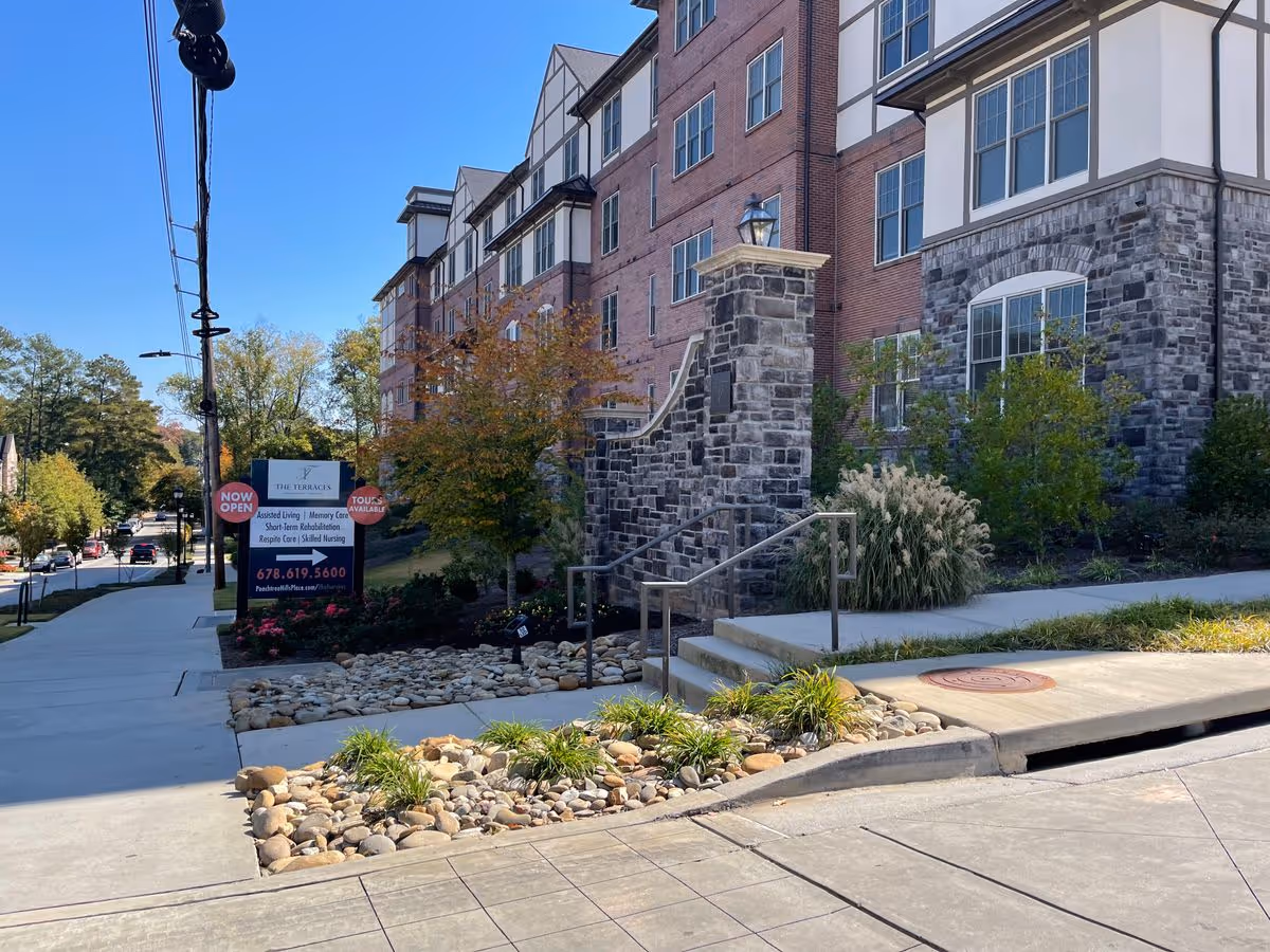 Street-level view of the brick-and-stone facade of The Terraces at Peachtree Hills Place with a sidewalk, landscaping, and an entrance sign.