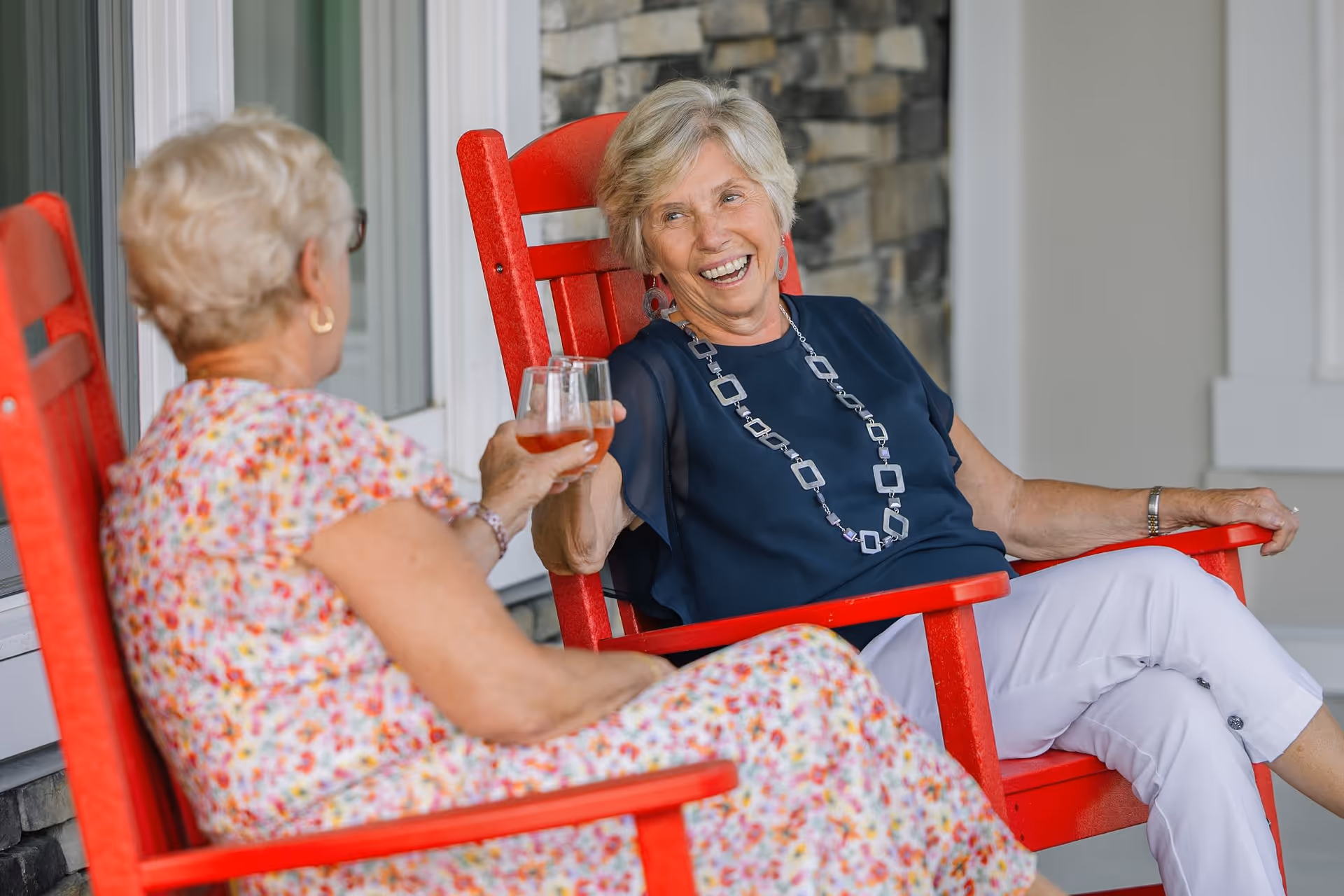 Two elderly women sitting on bright red wooden chairs on a porch, smiling and clinking glasses of a beverage, with a stone wall and white door in the background.
