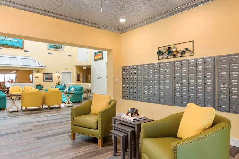 Interior view of a senior living facility lounge area with green and yellow armchairs, small nesting tables, and a wall of mailboxes. The room has light yellow walls and wood flooring. In the background, there are additional seating areas with teal and yellow chairs and tables, and some artwork on the walls.