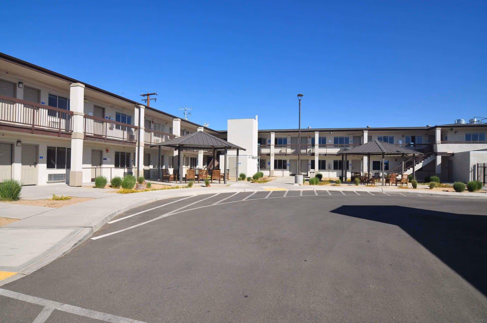 Two-story senior living building with an open courtyard, covered seating gazebos and a parking/drive area under a clear blue sky.