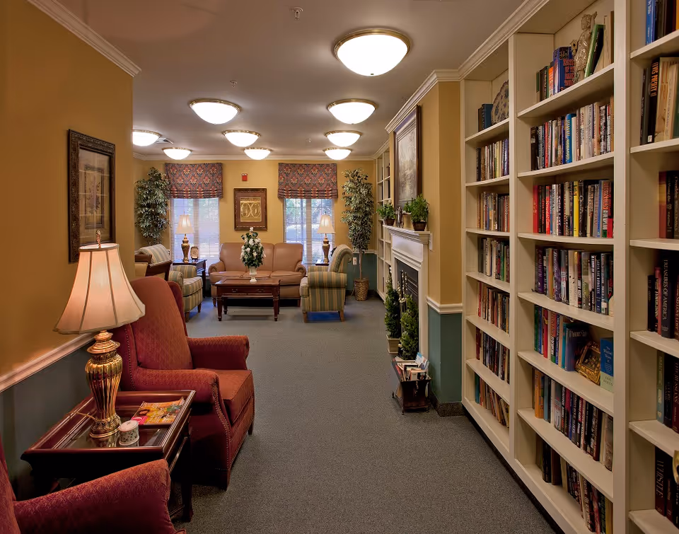A cozy living room area in a senior living facility featuring comfortable armchairs and sofas arranged around a wooden coffee table with a floral centerpiece. The room has built-in bookshelves filled with books on the right side, a fireplace with plants on the mantel, and framed artwork on the walls. The space is warmly lit with ceiling lights and table lamps, and windows with patterned valances allow natural light in.