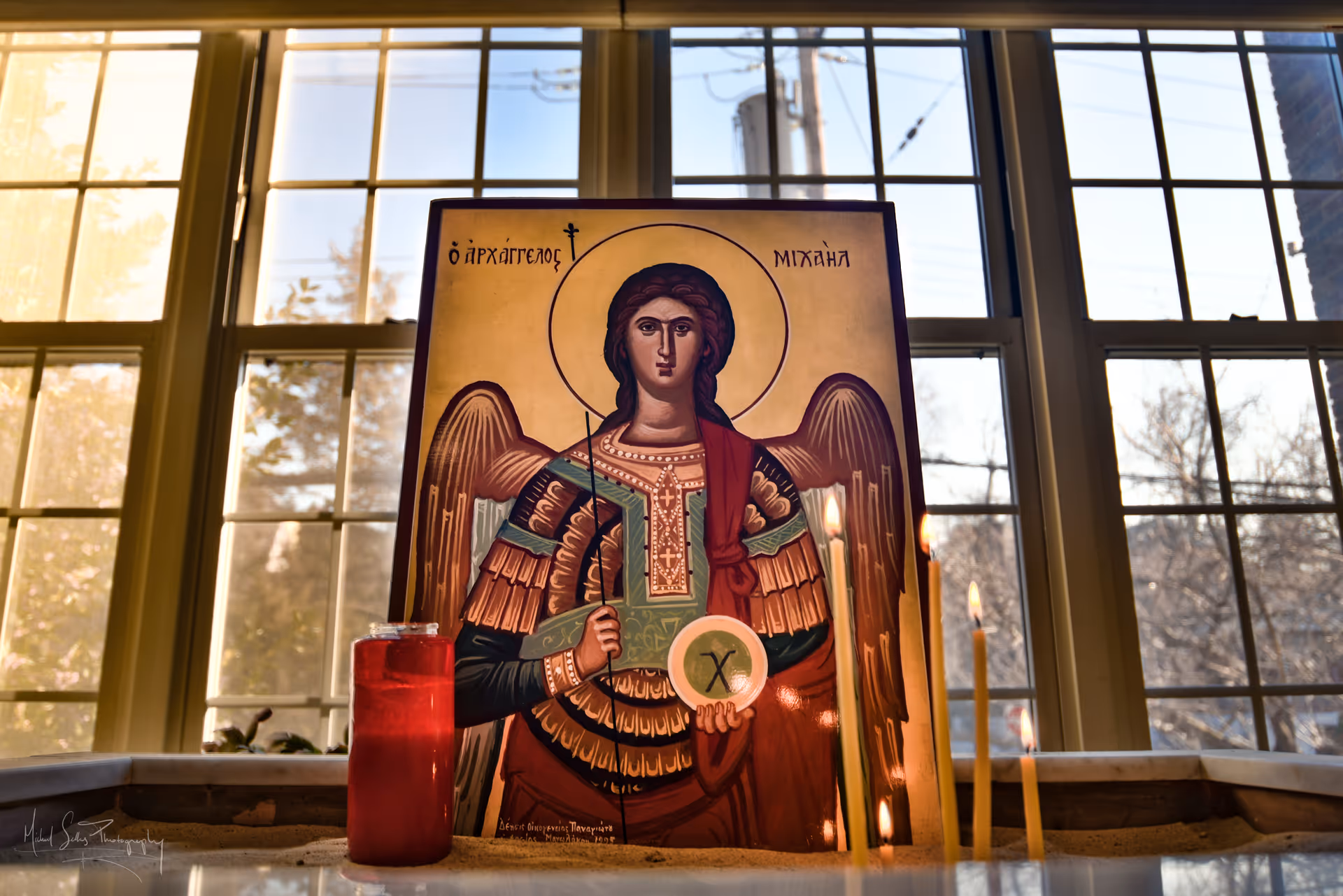 An icon painting of Archangel Michael displayed on a stand in front of a large window with multiple panes. The icon shows the archangel with wings, holding a staff and a circular object with a cross. In front of the icon, there is a red candle and several lit thin candles. The background outside the window shows trees and a utility pole.