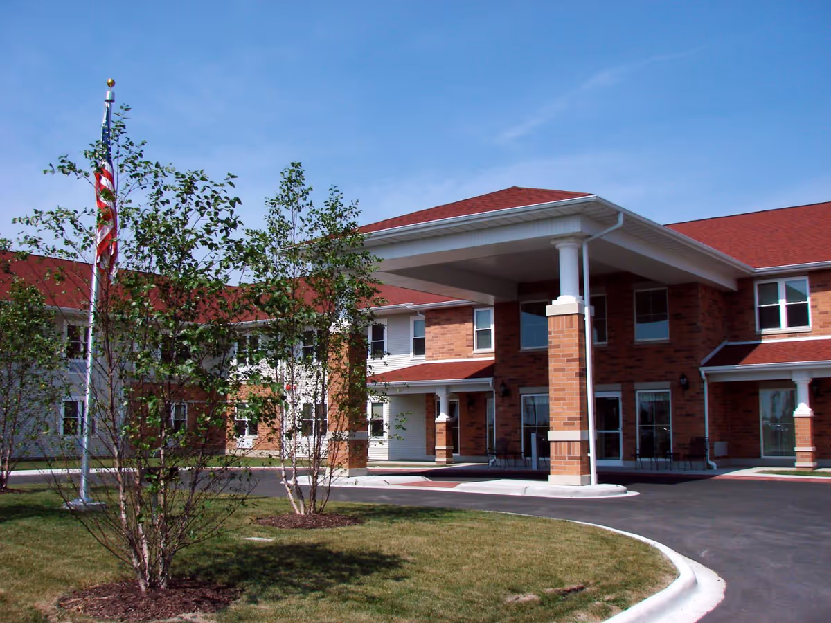 Exterior view of a senior living facility building with a covered entrance supported by columns, red roof, brick and siding walls, small trees, and an American flag on a flagpole in front.