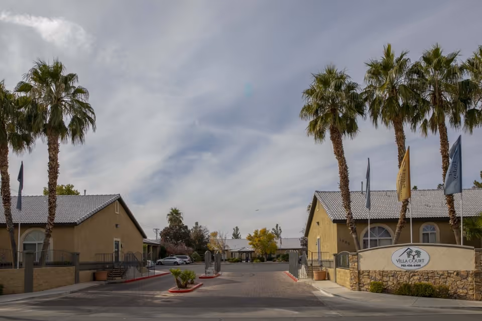 Entrance to Villa Court Assisted Living showing beige buildings, palm trees, flags, a driveway, and the facility sign at the front.