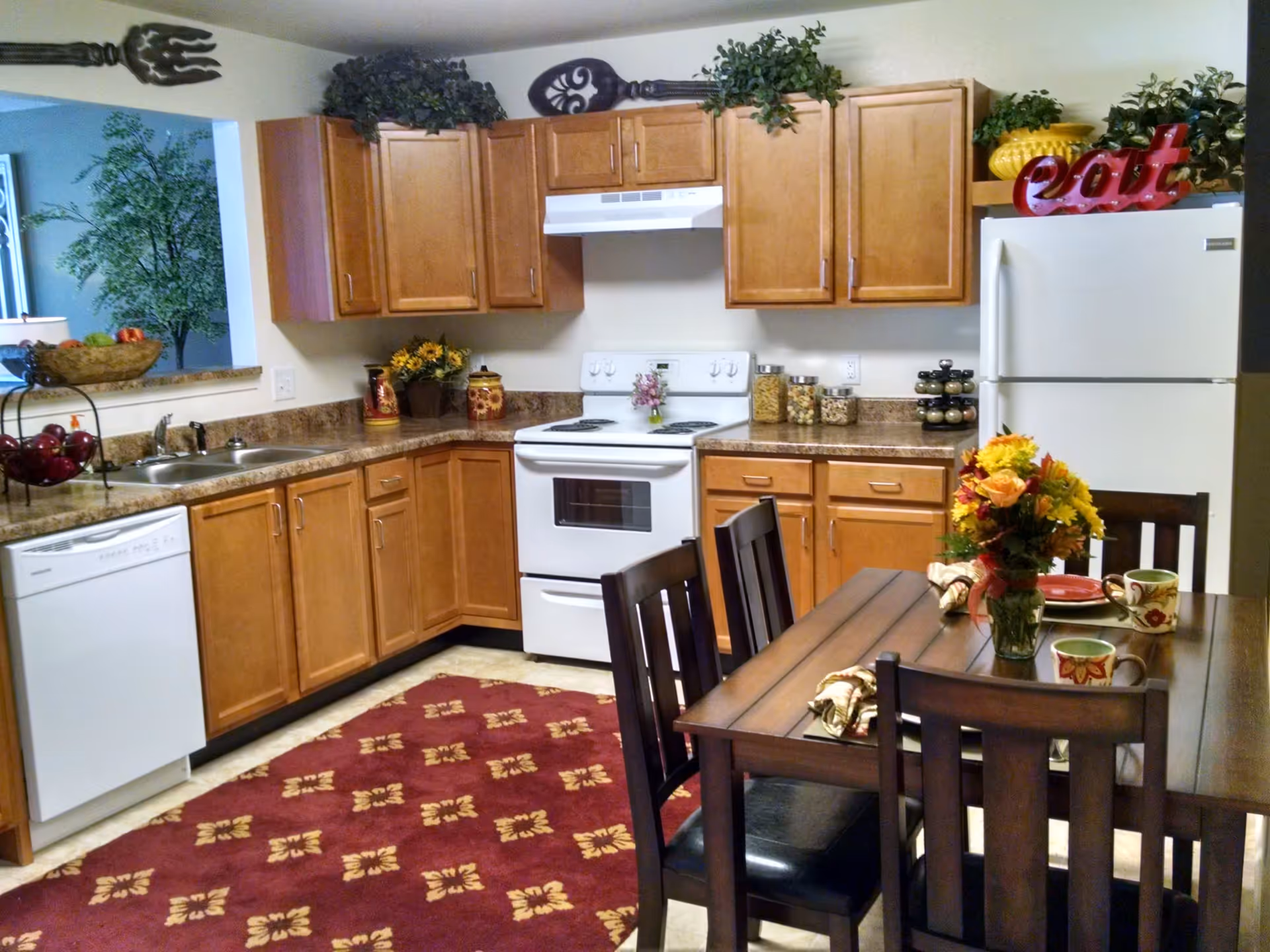 A cozy kitchen with wooden cabinets, a white stove and refrigerator, a dishwasher, and a double sink. The countertops have decorative jars and plants. A wooden dining table with four chairs is set with colorful mugs, plates, napkins, and a vase of flowers. The floor has a red patterned rug, and decorative oversized fork and spoon hang above the cabinets.