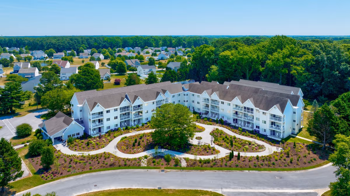 Aerial view of Gull Creek senior living facility showing a large, white, multi-story building with balconies surrounded by landscaped gardens and trees. The building is situated in a suburban area with many houses and dense forest in the background.