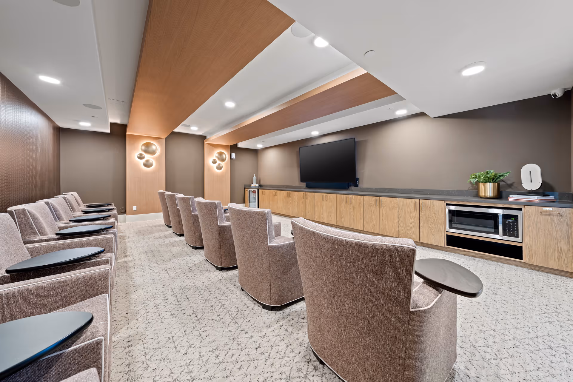 A modern media room with two rows of comfortable upholstered chairs equipped with small black side tables, facing a large flat-screen TV mounted on a brown wall. The room features a long wooden cabinet with a microwave, a small refrigerator, decorative items, and soft ceiling lighting with wooden panel accents.