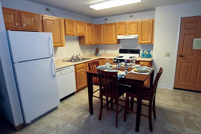 A kitchen with wooden cabinets, a white refrigerator, dishwasher, and stove. A wooden dining table set for four with plates, glasses, and napkins is in the center of the room. The floor is tiled, and there is a wooden door on the right side of the image.