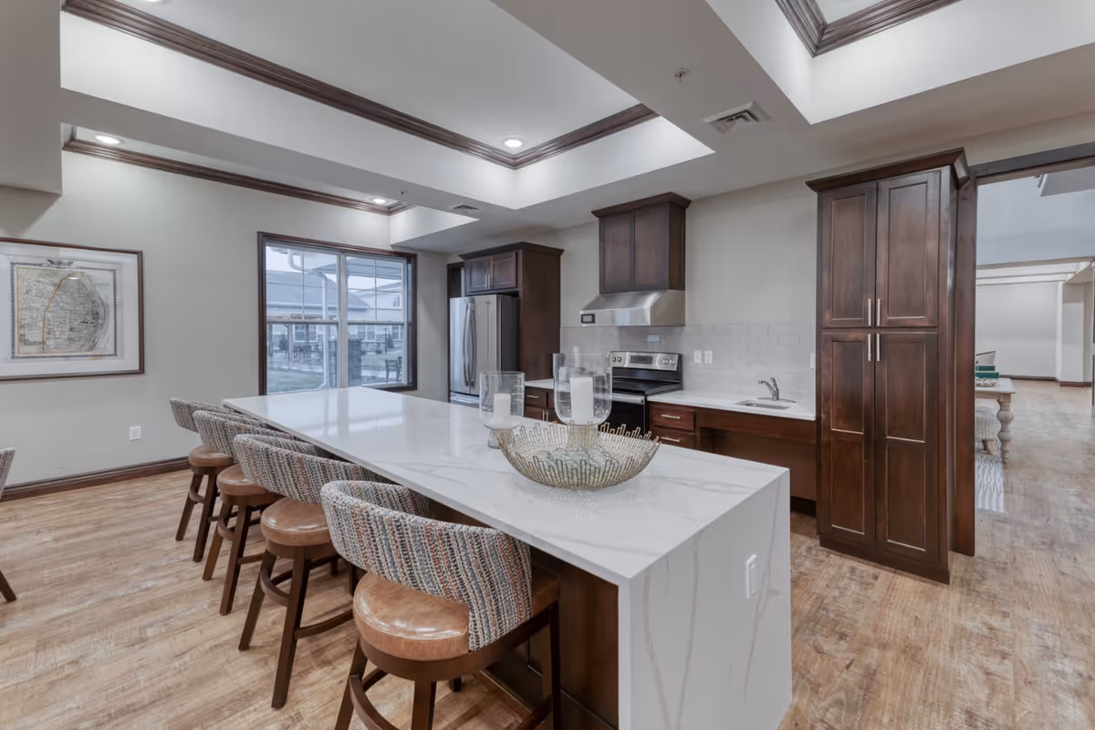 Modern kitchen area with a long white marble island countertop surrounded by six cushioned bar stools. The kitchen features dark wood cabinets, a stainless steel refrigerator, stove, and range hood. The room has wood flooring, recessed lighting, and a large window showing an outdoor view.