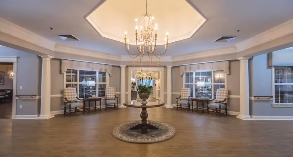 A spacious, elegant interior room with a round wooden table in the center holding a potted plant. The room features a chandelier hanging from a recessed ceiling, four cushioned armchairs with plaid upholstery arranged in pairs near the windows, and decorative columns along the walls. The floor is wooden, and the walls are painted light gray with white trim.