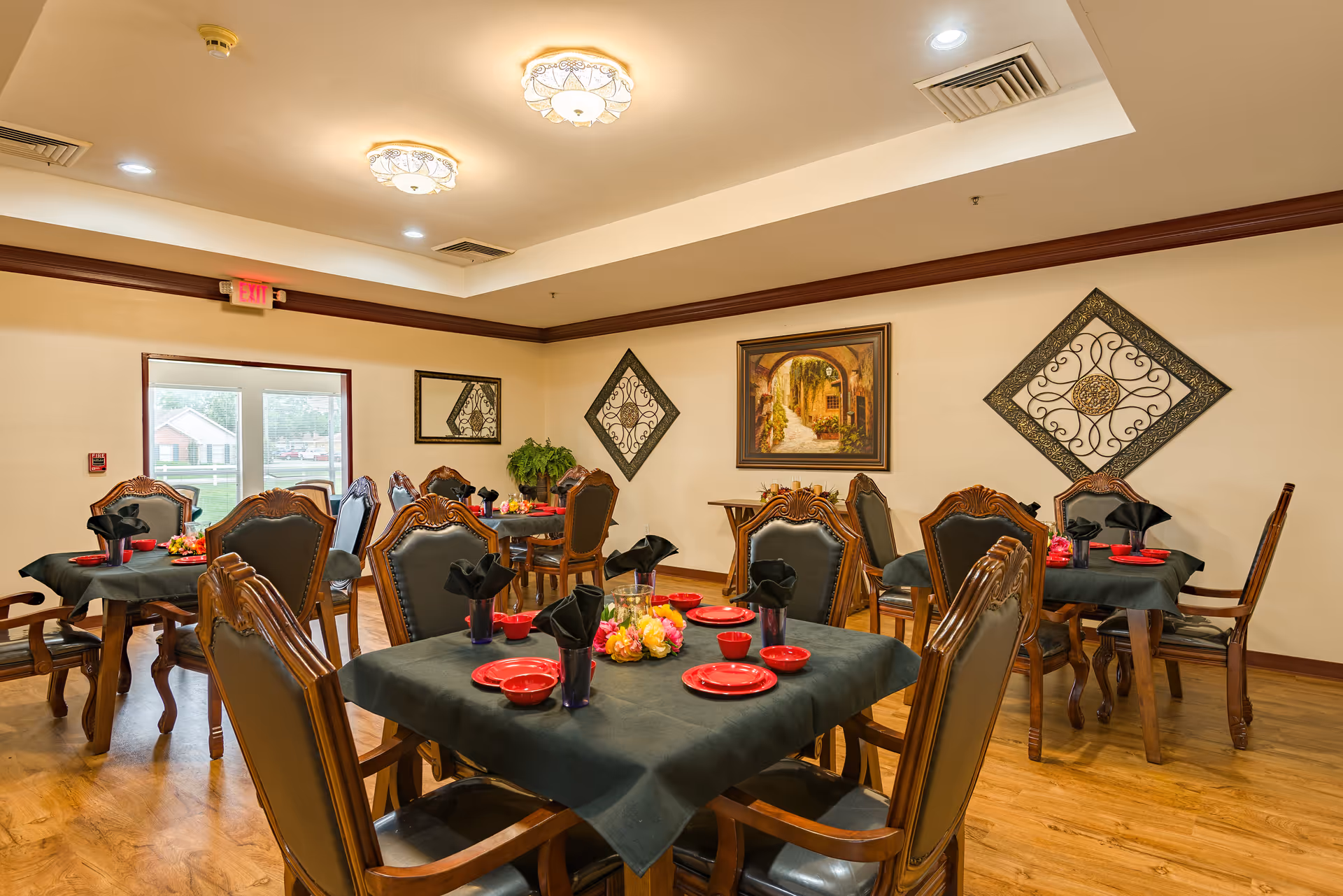 A dining room in Brentwood Retirement Community with multiple wooden tables covered with dark tablecloths. Each table is set with red plates, bowls, and black napkins in glasses. The room has wooden flooring, decorative wall art, and a painting of an archway with greenery. Ceiling lights illuminate the space, and there is a glass door leading outside.
