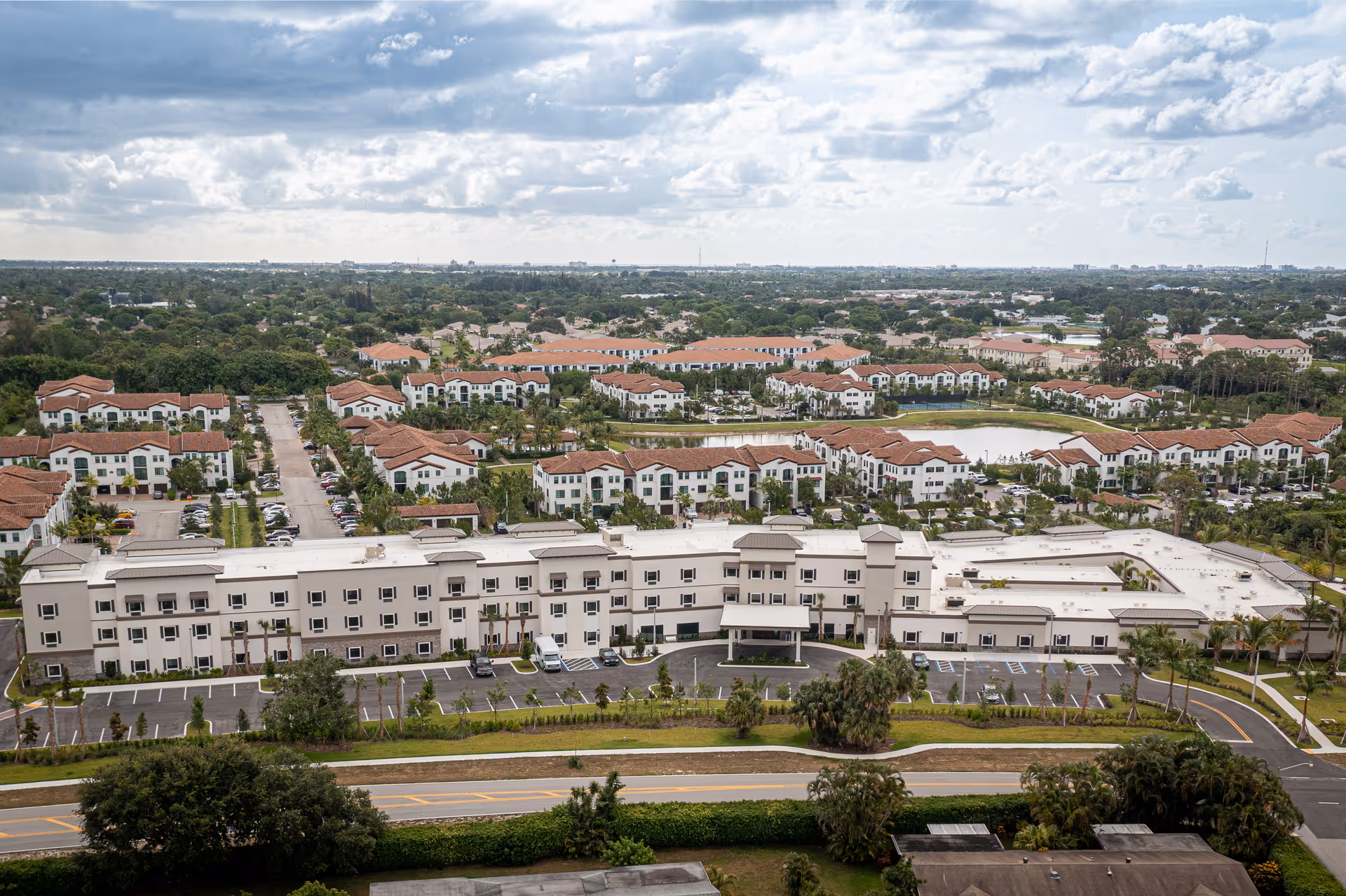 Aerial view of the Bella Mar Delray Beach senior living building and parking lot with surrounding residential neighborhood and cloudy sky.