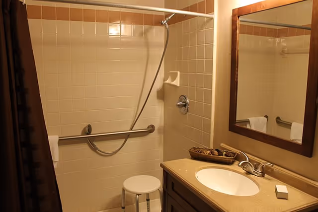 Accessible bathroom with tiled shower and grab bar, handheld showerhead and stool, and a vanity with sink and mirror.