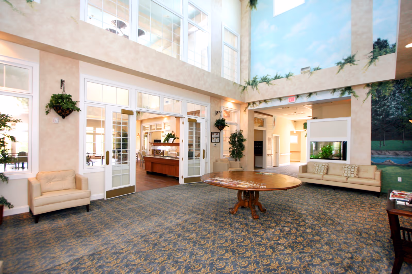 Spacious and well-lit common area in a senior living facility featuring high ceilings, large windows, and a patterned carpet. The room includes a round wooden table in the center, beige armchairs and a sofa along the walls, potted plants, and a mural of a natural landscape on one wall. Double glass doors lead to an adjoining dining area with wooden floors and additional seating.
