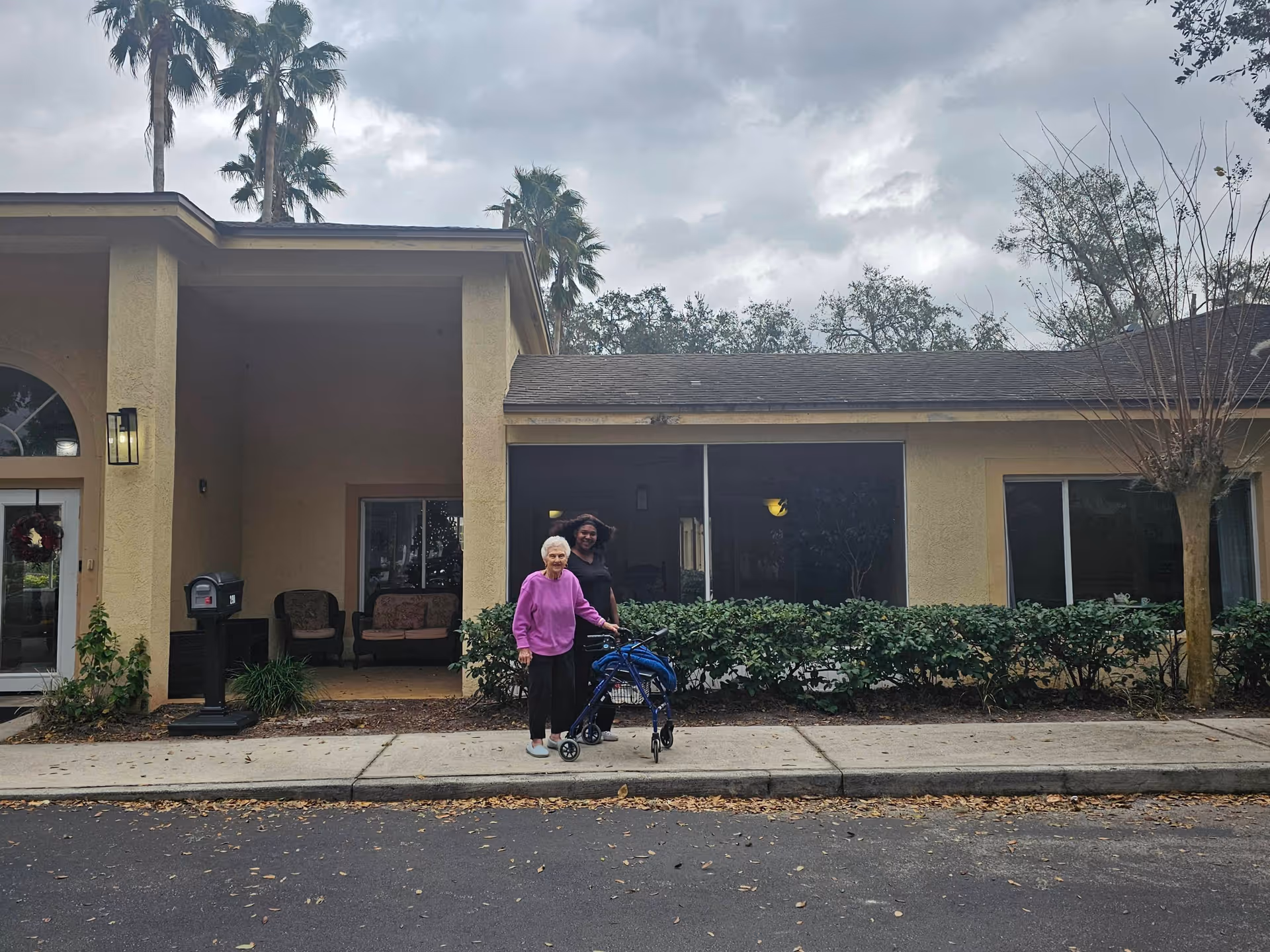 Two women standing outside a beige senior living facility building with large windows and a covered porch area. One woman is elderly and using a walker, and the other woman is standing behind her. There are palm trees and bushes around the building under a cloudy sky.