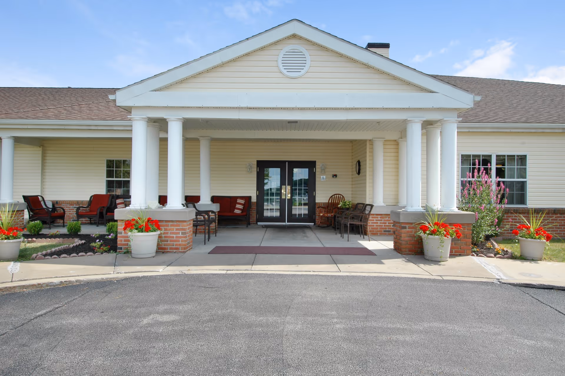 Front entrance of a single-story building with a covered porch supported by white columns. There are potted plants with red flowers on either side of the entrance, and outdoor seating with chairs and small tables is visible under the porch. The building has beige siding and a brick base with a double glass door entrance.