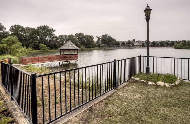 A lakeside view featuring a black metal fence enclosing a grassy area with a lamp post. Across the water, there is a wooden gazebo on a pier extending into the lake, surrounded by trees and distant buildings along the shoreline.