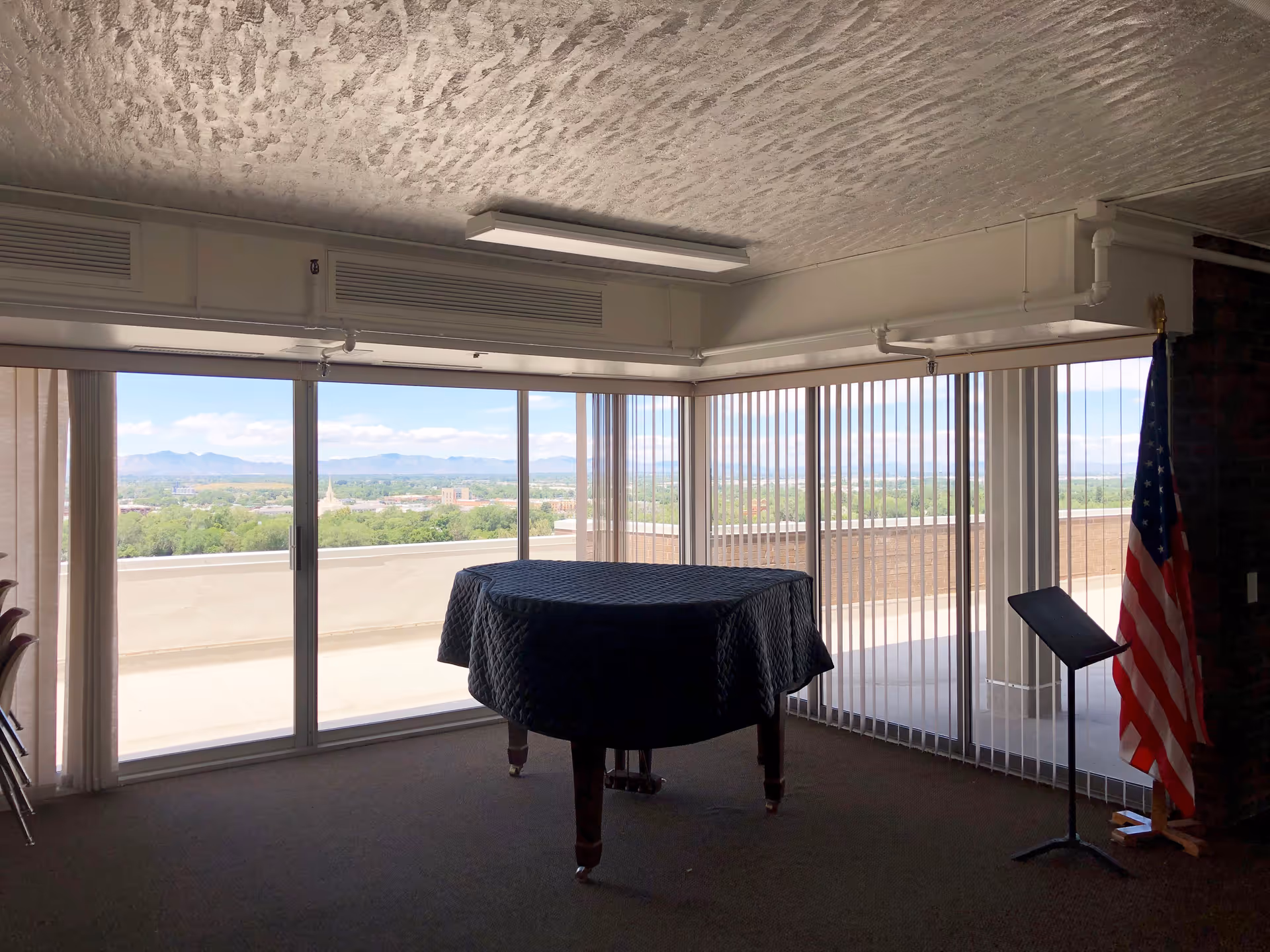 Interior room with large floor-to-ceiling windows and vertical blinds, showing a scenic view of distant mountains and greenery. A covered grand piano is centered in the room, and an American flag stands on a pole next to a music stand on the right side.