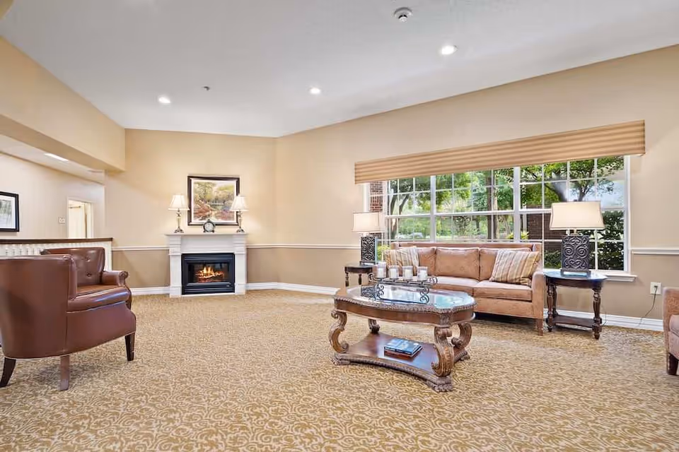 A cozy living room with a beige carpet and walls, featuring a brown leather armchair, a beige sofa with striped pillows, a wooden coffee table with candles and a book, two side tables with lamps, a fireplace with a painting above it, and a large window showing green trees outside.