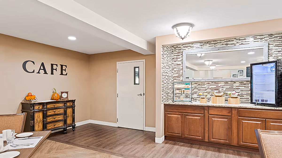 Interior cafe area with a serving counter, mirror, wooden cabinets, and a small buffet table under a wall sign reading 'CAFE'.