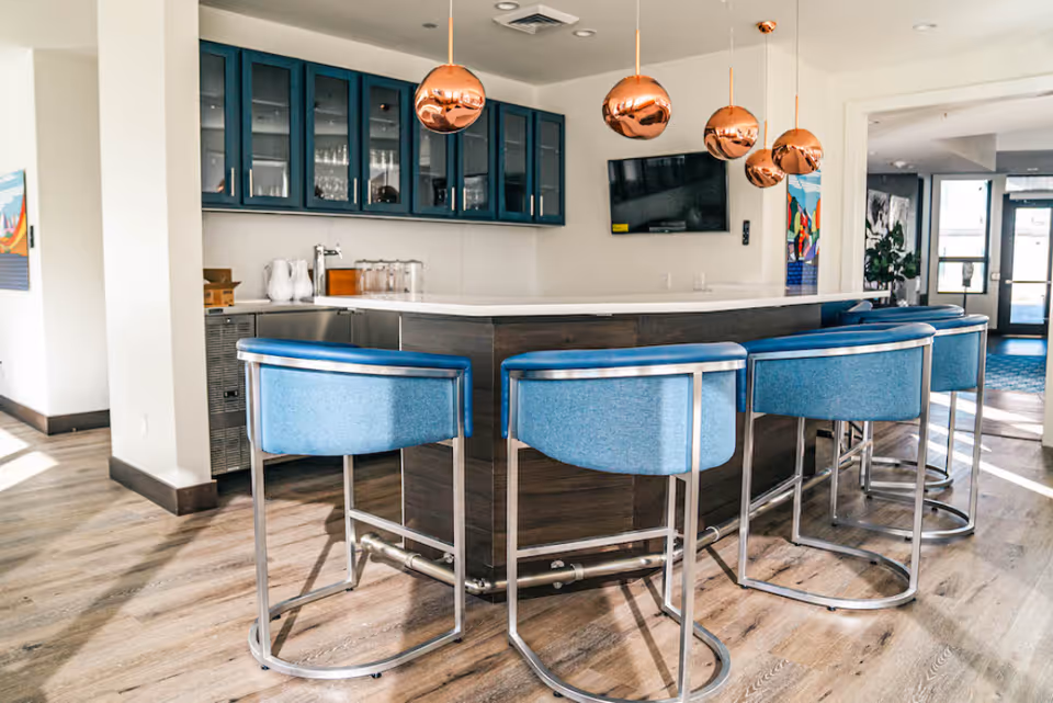 Bright common-area bar with blue upholstered stools around a white countertop, copper pendant lights, glass-front cabinets, and a wall-mounted TV.