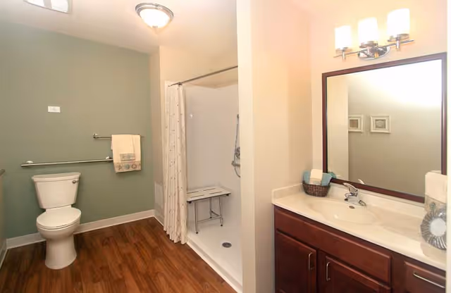 A clean and accessible bathroom featuring a white toilet, a walk-in shower with a white curtain and a foldable shower seat, a wooden vanity with a white countertop, a large mirror above the sink, and a towel rack with a beige towel. The walls are painted in light green and cream colors, and the floor has wood-style flooring.
