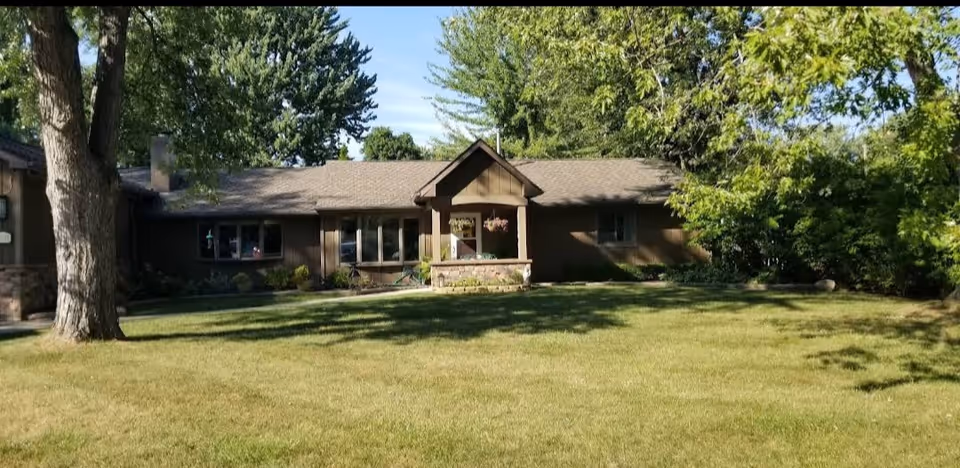 Single-story brown building with a covered front entrance, large grassy lawn, and surrounding trees.