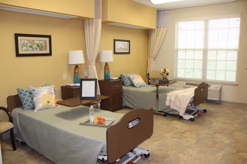 A bright and clean rehabilitation room with two hospital-style beds, each with a bedside table, lamps, and decorative pillows. The room has large windows with blinds, light yellow walls, and a beige floor. There are framed pictures on the walls and a small table with flowers near the window.