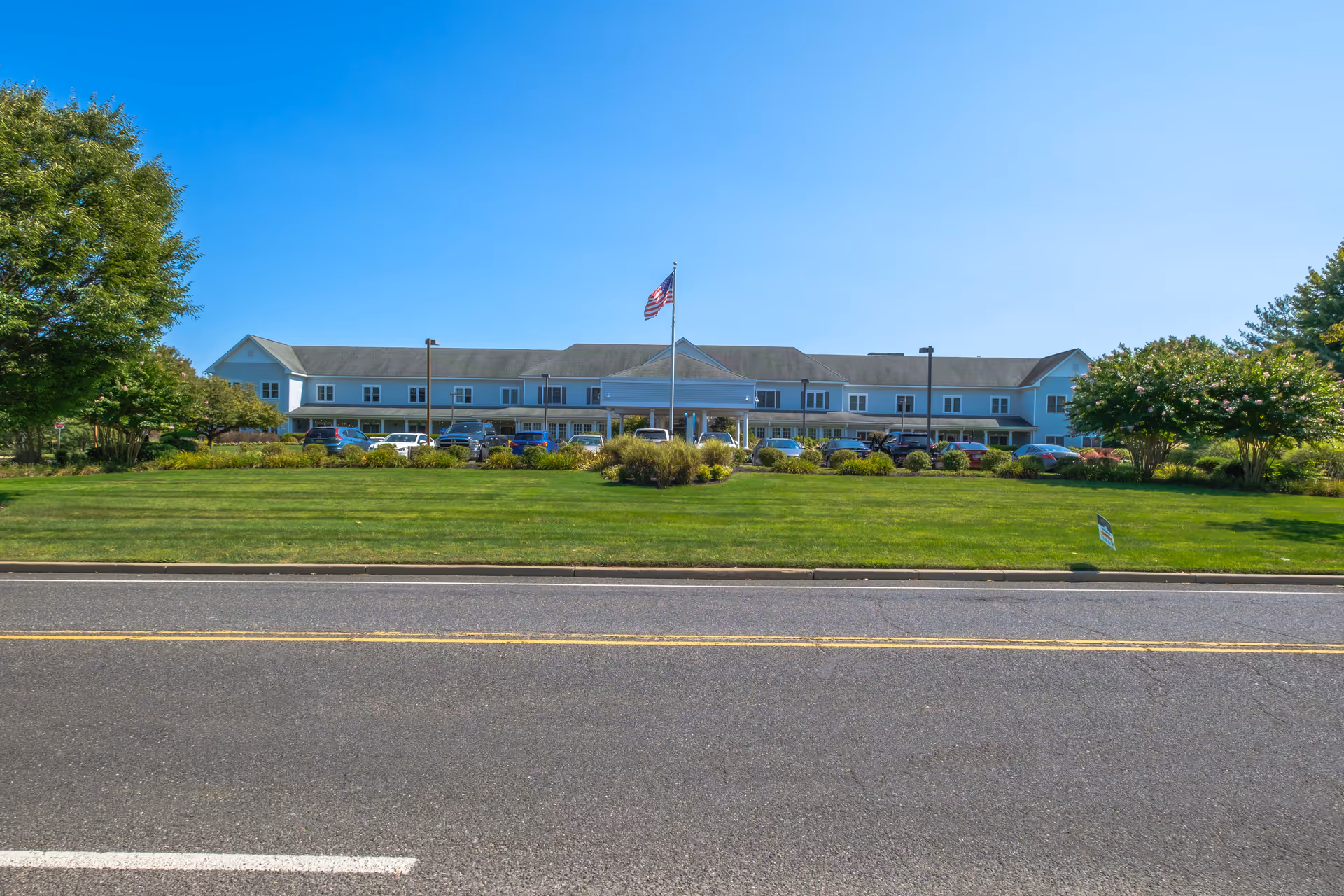 Front exterior view of a two-story senior living facility building with a large green lawn and an American flag on a flagpole in the center. Several cars are parked in front of the building under a clear blue sky.