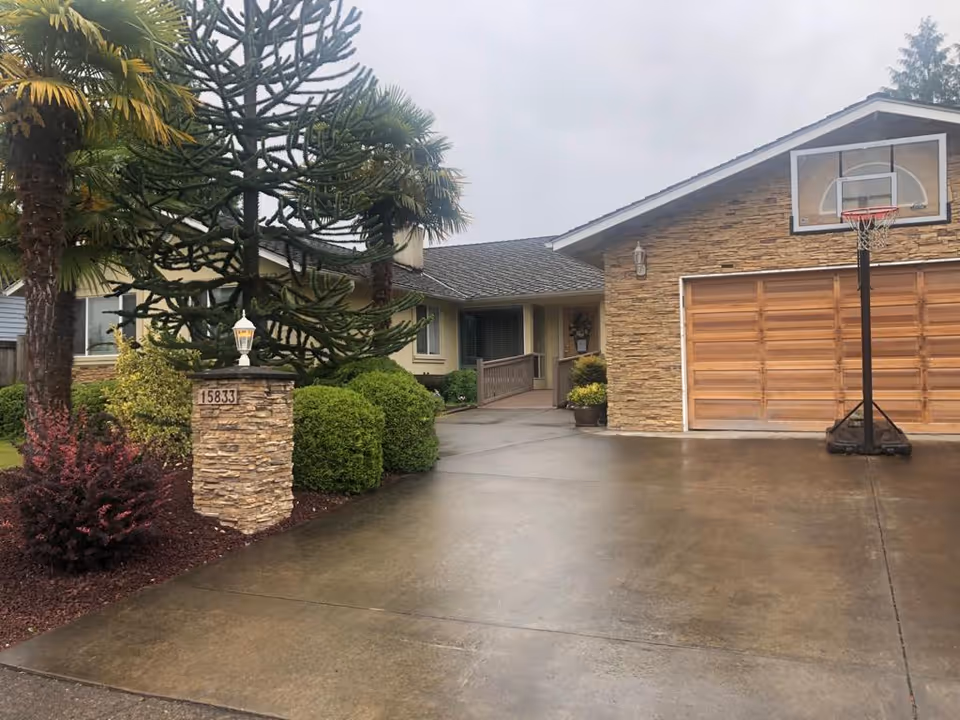 Exterior view of a single-story residential building with a stone facade garage and a basketball hoop on the driveway. The driveway is wet, and there are various shrubs and trees, including a palm tree, near the entrance. A stone pillar with the house number 15833 and a lantern is visible on the left side of the driveway.