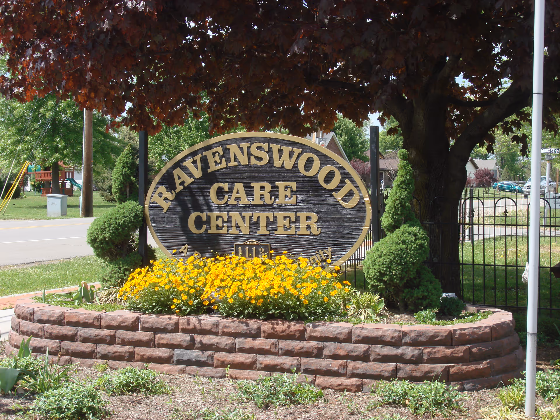 Wooden sign reading "RAVENSWOOD CARE CENTER" surrounded by yellow flowers and landscaping beneath a tree.