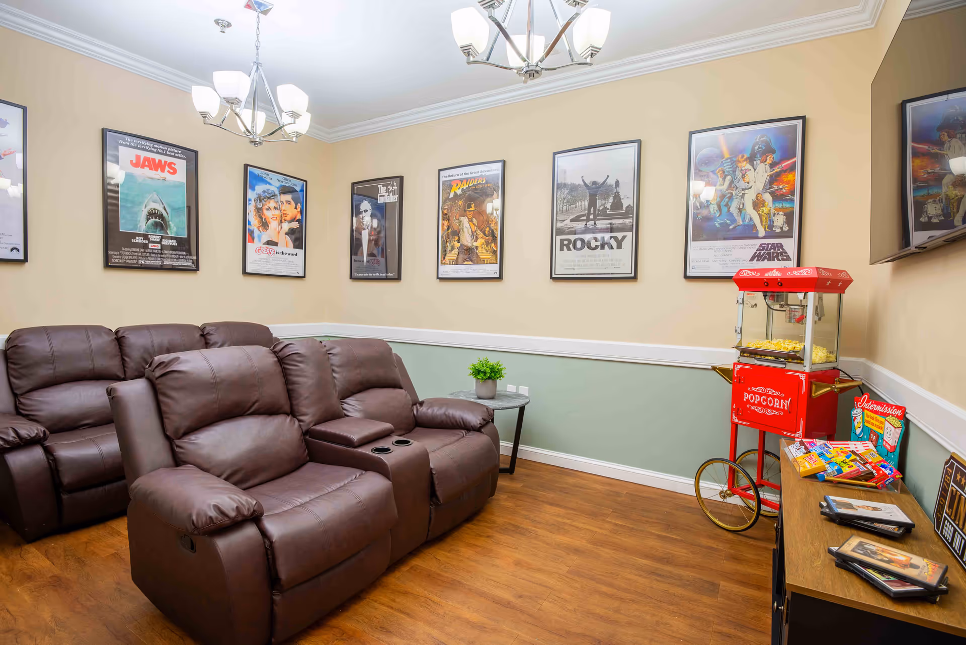 A cozy movie room with two rows of brown leather recliner seats facing a wall-mounted TV. The walls are decorated with framed classic movie posters including Jaws, Grease, The Godfather, Raiders of the Lost Ark, Rocky, and Star Wars. A red popcorn machine and a small table with snacks and DVDs are placed near the TV. The room has wooden flooring and soft yellow and green painted walls with white trim.